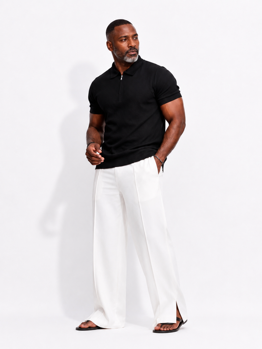 A man modeling a black short-sleeve polo shirt with a half-zip collar and white wide-leg trousers, standing against a plain white studio background.