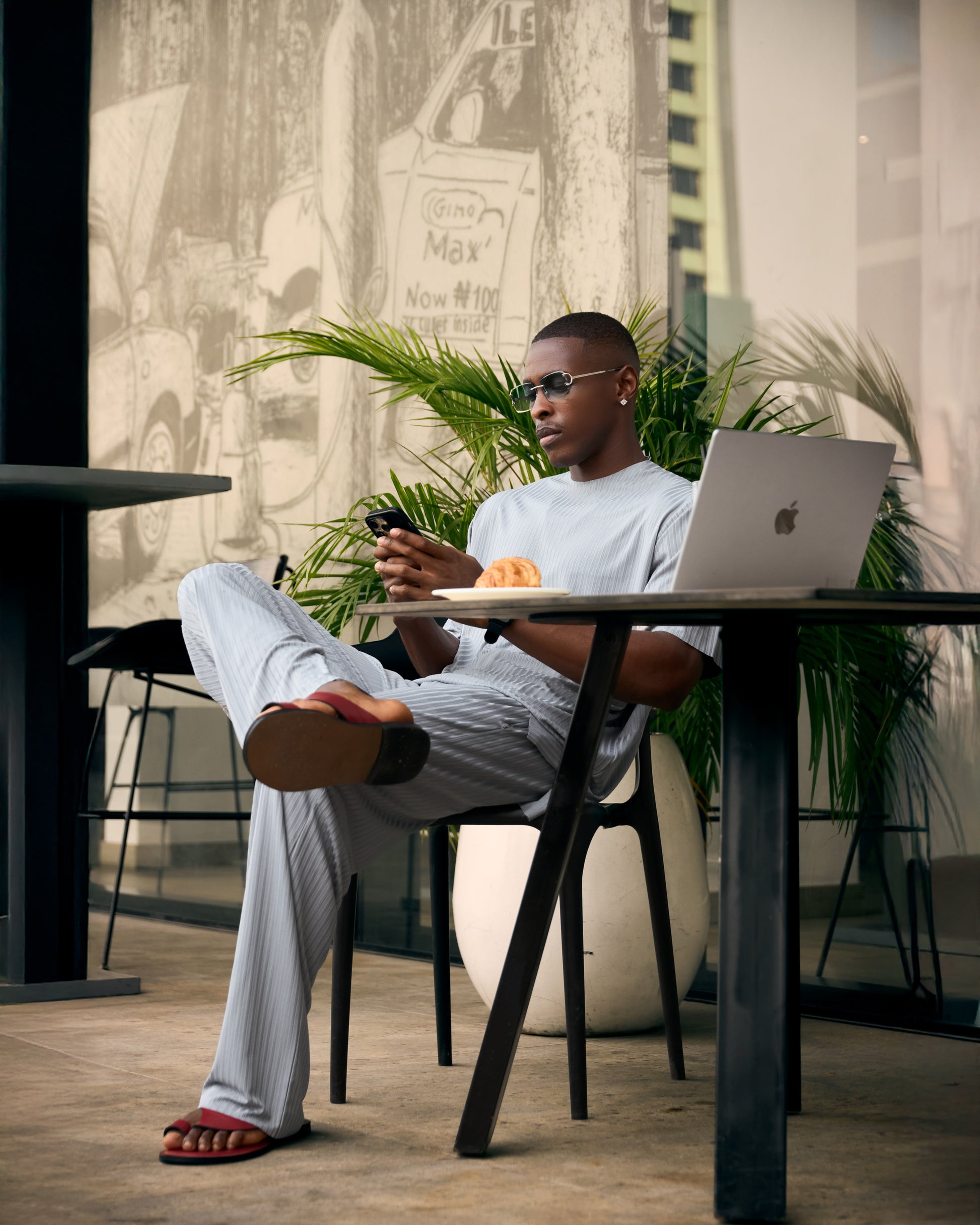 Man sitting at a table with a laptop and phone, surrounded by plants and cityscape.