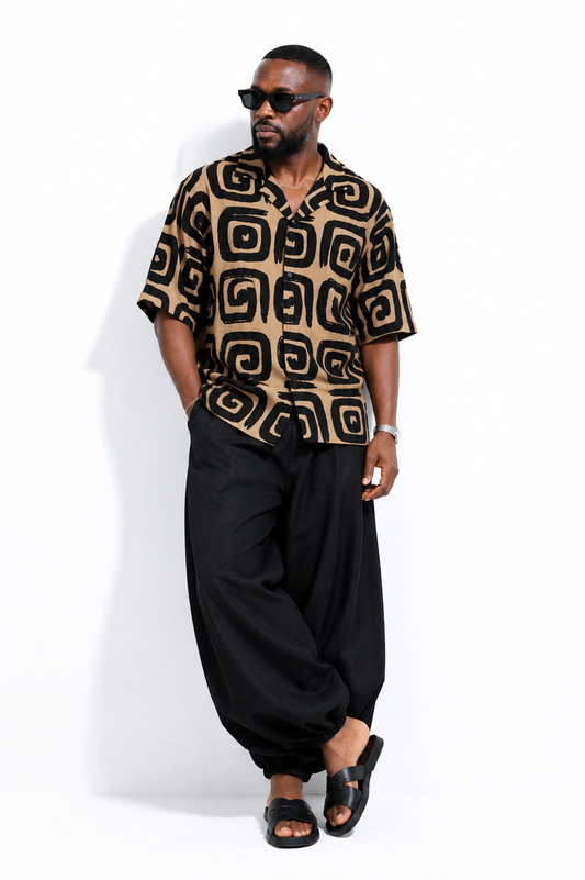 A man modeling a short-sleeve button-up shirt with a black and tan geometric pattern, paired with loose black pants and black slide sandals, set against a plain white studio background.