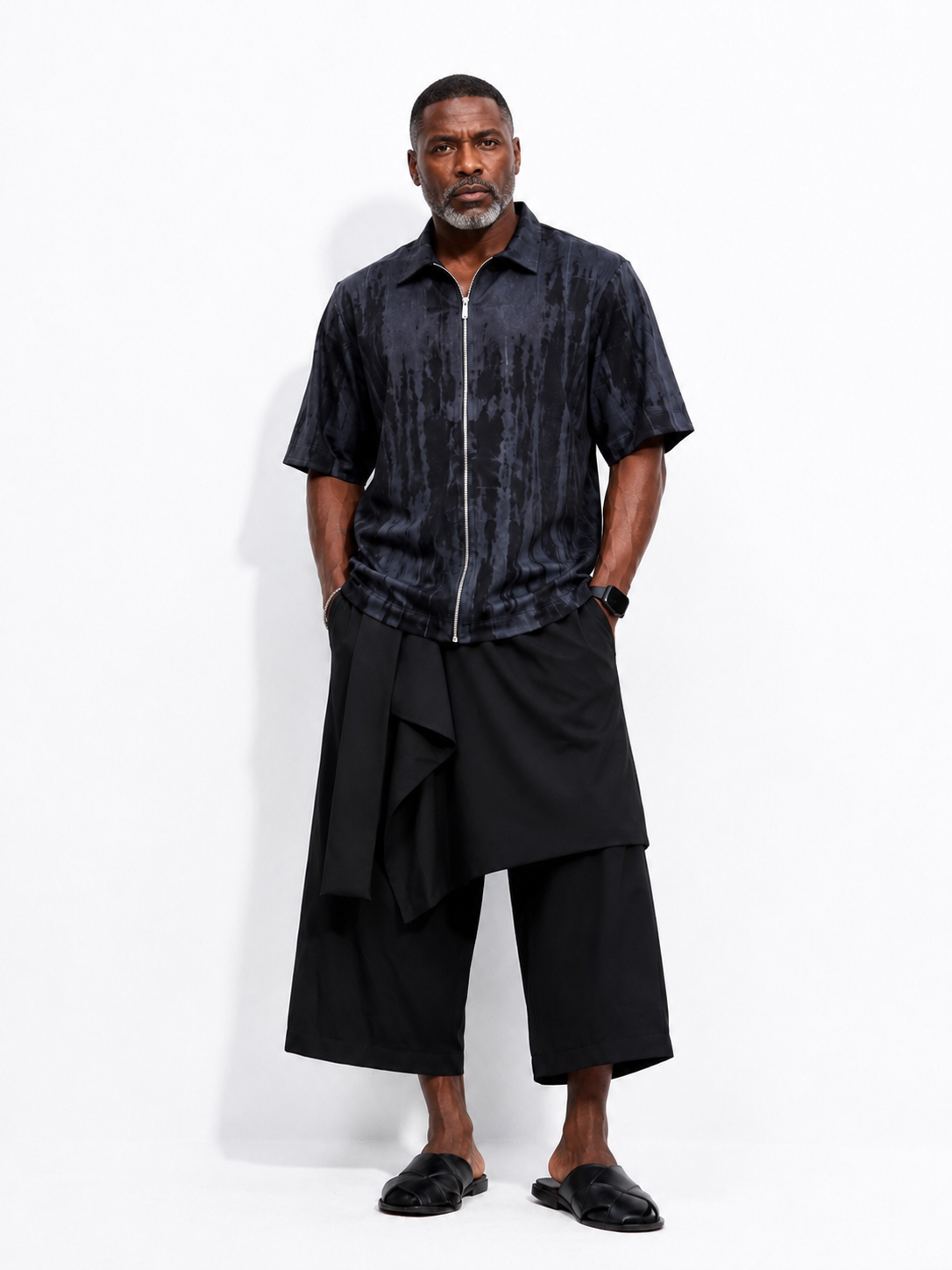 A man modeling a short-sleeve black button-up shirt with a vertical textured pattern and a front zipper, paired with black draped wide-leg pants and black slide sandals, against a plain white studio background.